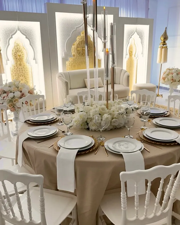 Table ronde nappée champagne, assiettes blanches, couverts dorés et chandeliers de verre devant le canapé des mariés